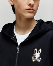 A person in a Psycho Bunny KIDS WADE CORD PATCH ZIP HOODIE (KT0500016)—a black premium cotton zip-hoodie with a cartoon bunny and crossbones chest logo—layered over a white t-shirt. Photo shows only the lower face and upper torso.