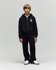 A young boy faces forward in a Psycho Bunny KIDS WADE CORD PATCH ZIP HOODIE (KT0500016) made from a premium cotton blend with a small cartoon graphic on the chest, paired with black cargo pants and white sneakers against a light gray background.