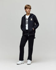 A person stands against a light background, wearing Psycho Bunny’s KIDS WADE CORD PATCH ZIP HOODIE (KT0500016) with an embroidered hood and bunny graphic, a matching white tee, black pants, and white sneakers, hands in their hoodie pockets.