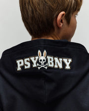A person in the Psycho Bunny KIDS WADE CORD PATCH ZIP HOODIE - KT0500016, a dark premium cotton blend zip-hoodie with "PSYBNY" and an embroidered bunny skull and crossbones logo on the back of the hood.