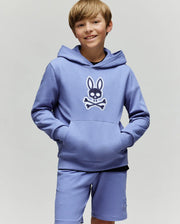A smiling boy with light brown hair wears the Psycho Bunny KIDS RYDER EMBROIDERED BUNNY PATCH HOODIE (KT0500018), featuring an embroidered Bunny patch and crossbones logo, standing against a plain light background.