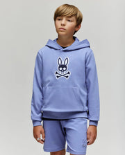 A young boy with light brown hair models the Psycho Bunny KIDS RYDER EMBROIDERED BUNNY PATCH HOODIE (KT0500018) and matching shorts, standing against a light gray background and gazing slightly to the side.