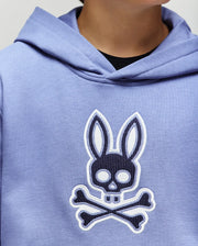 A person wears the Psycho Bunny KIDS RYDER EMBROIDERED BUNNY PATCH HOODIE (KT0500018) in light purple, featuring a large embroidered stylized skull and crossbones patch on the front. The focus is on this standout loungewear piece.