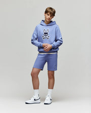 Teen boy stands against a plain background in a light blue Psycho Bunny KIDS RYDER EMBROIDERED BUNNY PATCH HOODIE (KT0500018), matching blue loungewear shorts, white socks, and white Nike sneakers.