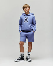A boy stands with hands in pockets, wearing the Psycho Bunny KIDS RYDER EMBROIDERED BUNNY PATCH HOODIE (KT0500018) in lavender—paired with matching shorts, black high-top sneakers, and striped socks against a light gray background.