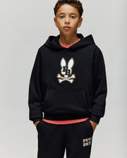 A boy stands against a plain background, wearing the Psycho Bunny KIDS WADE CORD PATCH HOODIE (KT0500031), featuring an embroidered Bunny and crossbones logo, with black shorts displaying "PSY" text on the left leg and his hands in his pockets.