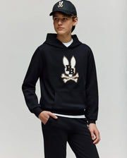 A teen boy wears the Psycho Bunny KIDS WADE CORD PATCH HOODIE (KT0500031), showcasing its premium cotton-blend fabric and signature Bunny patch with crossbones, paired with black pants and a black baseball cap.