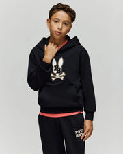 A boy wears the Psycho Bunny KIDS WADE CORD PATCH HOODIE (KT0500031) with an embroidered Bunny patch and matching black pants, standing against a plain light gray background.