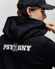 A person in the Psycho Bunny KIDS WADE CORD PATCH HOODIE (KT0500031), a black premium cotton blend, wearing a black cap and holding the hoodie near their neck. Features “PSYBNY” and bunny skull and crossbones graphic on the back.