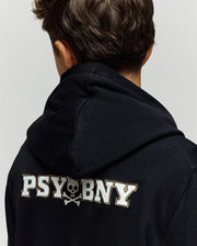 A child with short brown hair is seen from behind, wearing the Psycho Bunny KIDS WADE CORD PATCH HOODIE (KT0500031), a premium cotton blend black hoodie featuring "PSYBONY" and a small skull and crossbones in white on the back.