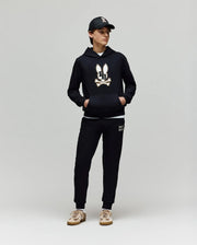 A person in a Psycho Bunny KIDS WADE CORD PATCH HOODIE (KT0500031), black joggers, black cap, and beige sneakers stands against a plain light gray background.