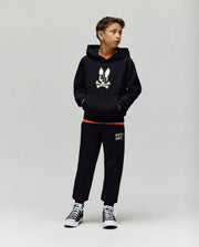 A boy stands against a plain background wearing the Psycho Bunny KIDS WADE CORD PATCH HOODIE (KT0500031), paired with black sweatpants featuring leg text and black high-top sneakers. He has his hands in his pockets and looks to the side.