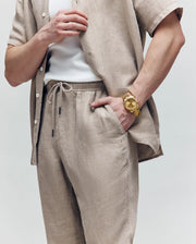 A person wears Psycho Bunny's MENS FARRIS WIDE LEG LINEN PANT in beige with a matching linen-blend shirt over a white tank top, accessorized with a large gold wristwatch; only the torso and arms are visible against a plain background.