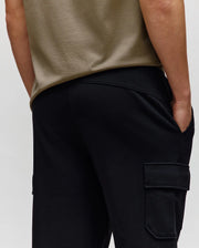 A person is shown from the back wearing a beige t-shirt and Psycho Bunny MENS NASH CARGO SWEATPANT - MB0800010, one hand in a pocket, highlighting both its utility cargo pockets and comfortable sweatpant style.