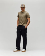 A man stands against a plain light gray background, wearing a khaki T-shirt, matching cap, white sneakers with brown soles, and Psycho Bunny MENS NASH CARGO SWEATPANT - MB0800010. His hands are in his pockets and he smiles slightly.