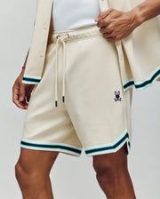 Wearing Psycho Bunny's MENS BASIL TAPED SHORT - MB0900005 in a soft cotton-modal blend with green, blue, and black trim and a bunny skull logo on the left leg, paired with a matching open shirt over a white top for classic confidence.