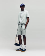 A man stands against a plain background wearing Psycho Bunny MENS ROBERT KNIT STRIPE SHORT - MB0900007 with a matching shirt, graphic tee, blue cap, white socks, dark sneakers, sunglasses, and carrying a beige tote bag.
