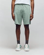 A person faces forward against a plain light background wearing Psycho Bunny's MENS RON WAFFLE CARGO SHORT - MB0900012 in sage green, paired with a white T-shirt, gray socks, white and green sneakers, and a bracelet.