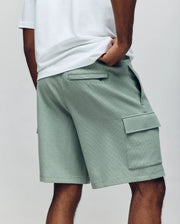A person is seen from the back wearing Psycho Bunny MENS RON WAFFLE CARGO SHORT - MB0900012 in sage green with flap pockets and visible stitching, paired with a white t-shirt and one hand in their pocket.