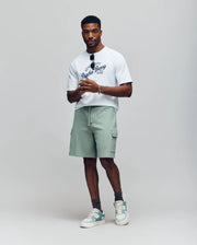 A man stands against a neutral background wearing a white graphic t-shirt, grey socks, pale green and white sneakers, holding sunglasses, and Psycho Bunny MENS RON WAFFLE CARGO SHORT - MB0900012 in light green with drawstring and pockets.