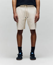 A person wears Psycho Bunny MENS KAI TWILL TEXTURE SWEATSHORT - MB0900015 in beige, featuring a twill texture and embroidered Bunny logo, with black socks, black sneakers, and a black shirt. Only the lower half is visible against a light gray background.
