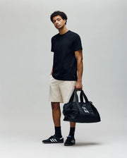 A young man stands against a plain background wearing a black Psycho Bunny t-shirt, MENS KAI TWILL TEXTURE SWEATSHORT - MB0900015, black sneakers and socks. He holds a large black duffel bag in his left hand, right hand in his pocket, looking relaxed.