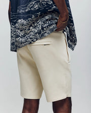 A person wears Psycho Bunny’s MENS LAZSLO SWEATSHORT - MB0900016 in cream, made from a modal-cotton blend with side and back pockets, paired with a dark patterned shirt. Only the lower torso and upper legs are shown—ideal for casual style.
