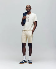 A man poses against a plain background in a beige bunny-graphic T-shirt, Psycho Bunny MENS LAZSLO SWEATSHORT - MB0900016, black sneakers with white socks, and holds a black jacket over his shoulder for a cool, casual look.