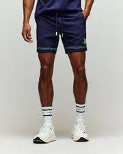 Wearing Psycho Bunny’s TENNIS BALL CREW MENS SHORT - MB0900017 in navy with neon green accents, a matching top, white crew socks with black text, and white sneakers, a person stands against a plain light background—ready for tennis or training.