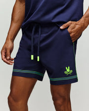 A person wears Psycho Bunny’s TENNIS BALL CREW MENS SHORT (MB0900017) in navy, featuring neon green accents, a drawstring, and a crossed rackets bunny logo graphic on the left leg. Their hand is in the right pocket.