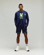 A man stands against a plain background wearing Psycho Bunny’s TENNIS BALL CREW MENS SHORT - MB0900017 in navy blue, paired with a tennis-themed graphic tee, quick-dry jacket, white sneakers, and striped socks.