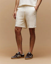Against a beige background, only the lower body is visible, wearing Psycho Bunny MENS KAIRO CROCHET SHORT - MB0900026 in cream with a matching top and black Adidas sneakers with white stripes; hands relaxed by their sides—a perfect summer look.