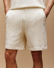Close-up of a person wearing Psycho Bunny MENS KAIRO CROCHET SHORT - MB0900026 in textured off-white, with hands in pockets, cotton drawstring waist, and small embroidered logo on the left leg against a beige background.