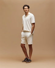 A man stands against a tan background, wearing Psycho Bunny's MENS KAIRO CROCHET SHORT - MB0900026 in cream with a short-sleeve shirt and brown sneakers. One hand is in his pocket.