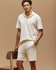 A man stands beside a wooden chair, wearing Psycho Bunny’s MENS KAIRO CROCHET SHORT (MB0900026) and a matching cream crochet polo, hand in pocket against a beige background—his ideal summer uniform.