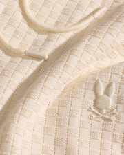 Close-up of Psycho Bunny’s MENS KAIRO CROCHET SHORT (MB0900026) in cream-colored, checkered knit with a raised bunny and crossbones logo. Features a cream drawstring with plastic tips—ideal for summer wear.