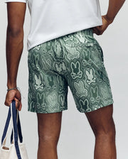 A person seen from behind wears Psycho Bunny MENS CHUCK ALL OVER PRINT LIGHTWEIGHT SWIM TRUNK (MB1000003)—green eco-friendly shorts with abstract bunny patterns—holding a tote in their left hand, emphasizing the sustainable design.