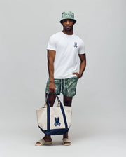 A man stands against a plain background wearing a white t-shirt, Psycho Bunny MENS CHUCK ALL OVER PRINT LIGHTWEIGHT SWIM TRUNK in green recycled polyester, a matching bucket hat, beige slides, and holds a beige and navy tote bag with the blue Psycho Bunny logo.