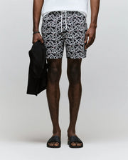 A person wears Psycho Bunny MENS CYRUS ALL OVER PRINT SWIM TRUNK - MB1000004 (black and white pattern), a white shirt, black sandals, and holds a black garment. The plain light background does not reveal the person’s face.