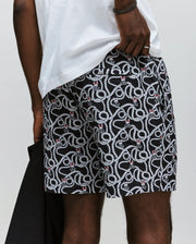 A man wears Psycho Bunny's MENS CYRUS ALL OVER PRINT SWIM TRUNK - MB1000004, featuring a red heart pattern and white rope design, paired with a white T-shirt and dark jacket; only his lower torso and upper legs are shown.