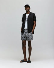 A man stands against a plain background wearing a black cap, Psycho Bunny MENS CYRUS ALL OVER PRINT SWIM TRUNK - MB1000004, a short-sleeve black shirt, graphic white tee, and black slide sandals. His hands are in his pockets.