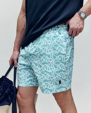 A person wears Psycho Bunny MENS CYRUS ALL OVER PRINT SWIM TRUNK - MB1000004 in aqua blue/white, a navy shirt, a wristwatch, and holds a beige tote with a navy towel. Only their torso and legs are visible.