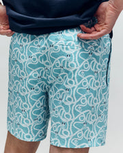 A person wears Psycho Bunny’s MENS CYRUS ALL OVER PRINT SWIM TRUNK (MB1000004) in nautical light blue, featuring white rope and smiling snake face patterns. The quick-dry seersucker fabric adds comfort as their hands rest behind their back.