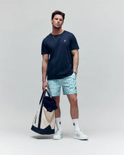 A man poses casually against a plain background wearing a navy t-shirt, Psycho Bunny MENS CYRUS ALL OVER PRINT SWIM TRUNK - MB1000004, white socks, white sneakers, and holding a large tote bag.
