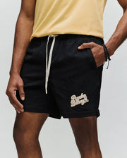A person in a light yellow t-shirt and Psycho Bunny MENS ELIAS SWIM TRUNK (MB1000008)—black, textured, with white drawstring, no-chafe netting, and Psycho Bunny logo on the left leg—stands with one hand in their pocket.