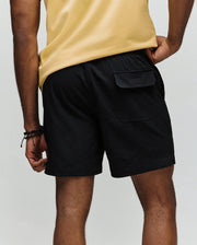 A person in a yellow shirt and Psycho Bunny MENS ELIAS SWIM TRUNK - MB1000008, classic fit and black color, stands with their back to the camera, showing a right pocket flap. Their left hand is on their hip; they wear black bracelets.