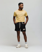 A man stands against a plain background wearing a yellow Psycho Bunny t-shirt, MENS ELIAS SWIM TRUNK - MB1000008 with classic fit and no-chafe netting, white striped socks, beige sneakers, and sunglasses hanging from his shorts.