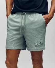 Wearing Psycho Bunny's MENS ELIAS SWIM TRUNK - MB1000008 in light sage green with textured fabric, no-chafe netting, drawstring, and the Psycho Bunny logo on the left leg, a person stands hands-in-pockets with a black tee showing.