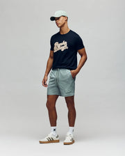 A man in a light green cap, black graphic t-shirt, Psycho Bunny MENS ELIAS SWIM TRUNK - MB1000008 with classic fit and no-chafe netting, white socks, and white sneakers stands against a plain light background.
