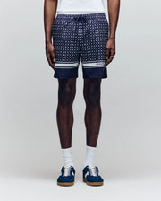A man wears Psycho Bunny MENS HUNTER ALL OVER PRINT SWIM TRUNK (MB1000011), featuring a navy geometric pattern, quick-dry fabric, drawstring waist, white hem stripes, paired with white socks and blue sneakers against a plain light background.